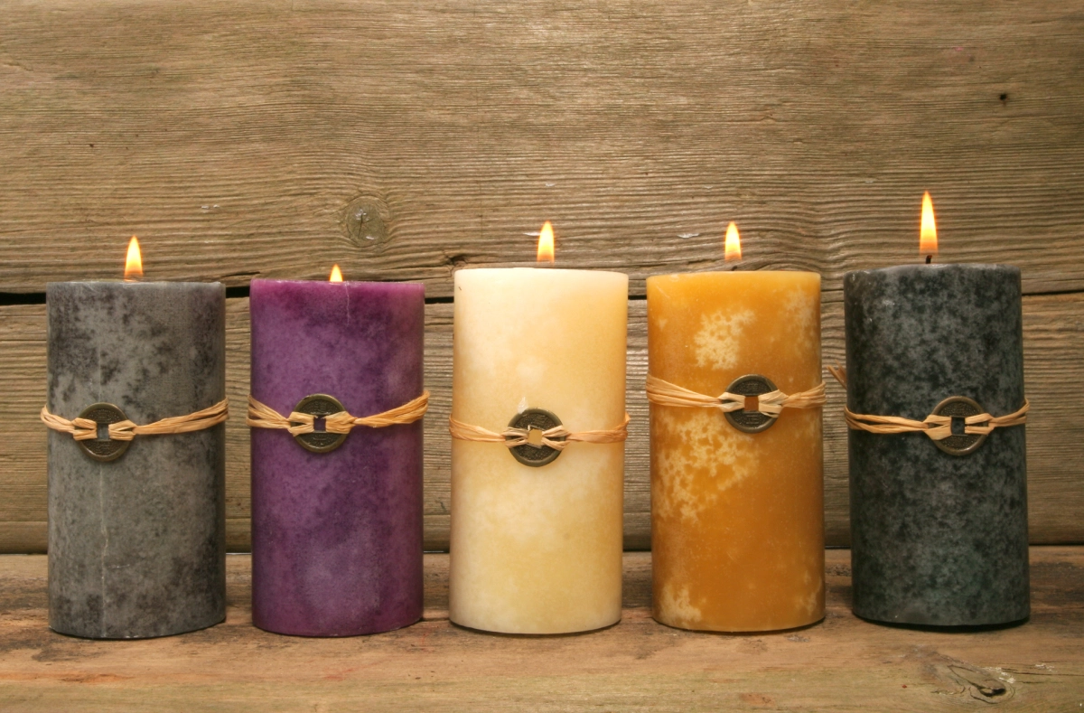 Five lit candles in gray, purple, cream, yellow, and dark gray stand in a row on a wooden surface. Each has a rustic twine and metal adornment. Background is a wooden panel.