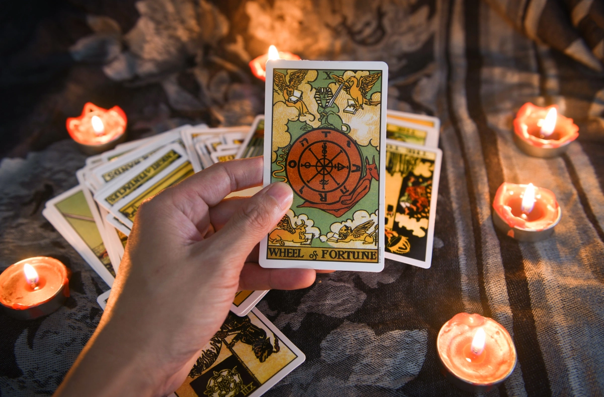 Hand holding a "Wheel of Fortune" tarot card surrounded by lit candles and other tarot cards on a dark surface.