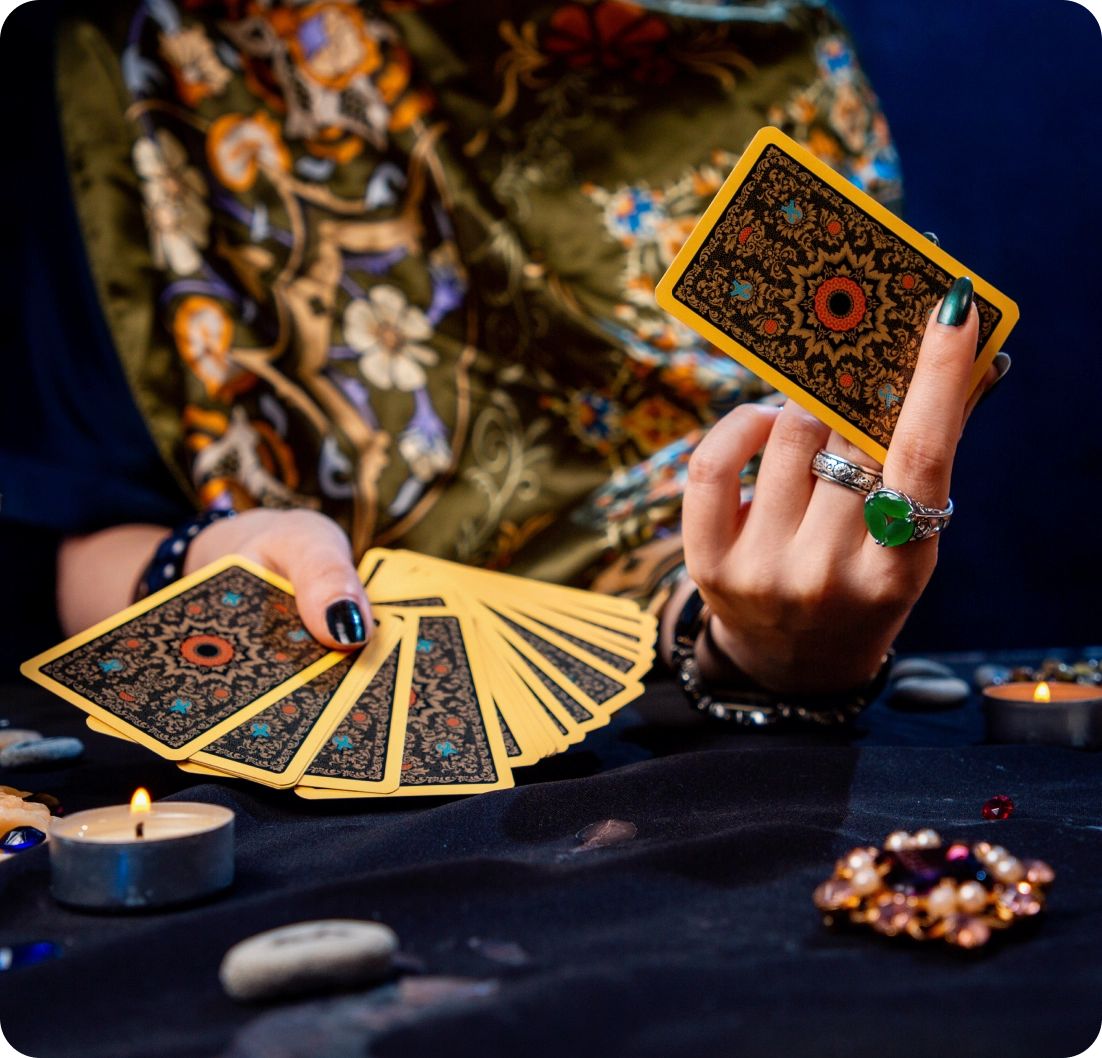 Person holding tarot cards, wearing rings and a patterned scarf. Cards are fanned out on a dark cloth with a lit candle and stones.