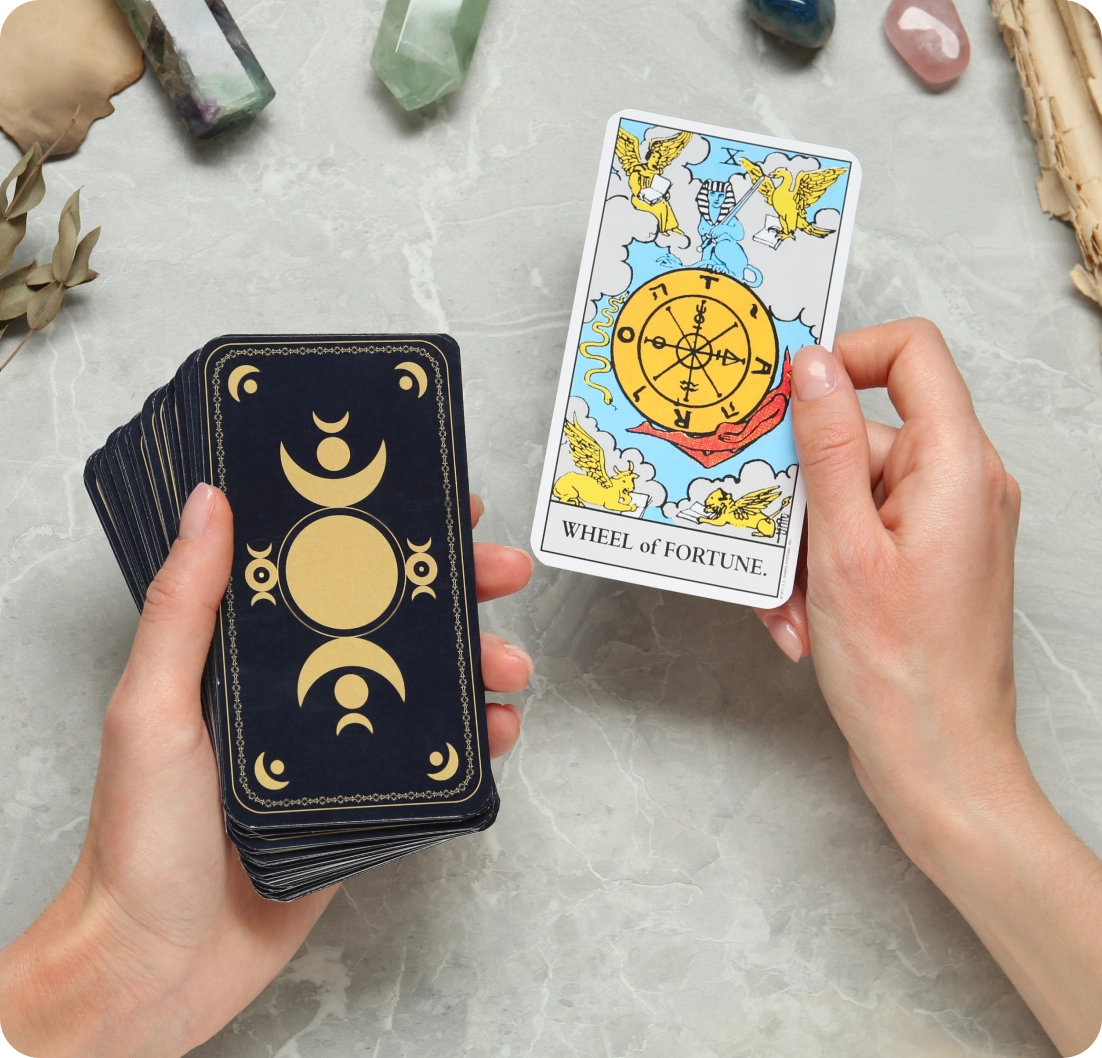 Hands holding a tarot card deck and displaying the Wheel of Fortune card on a marble surface with crystals nearby.