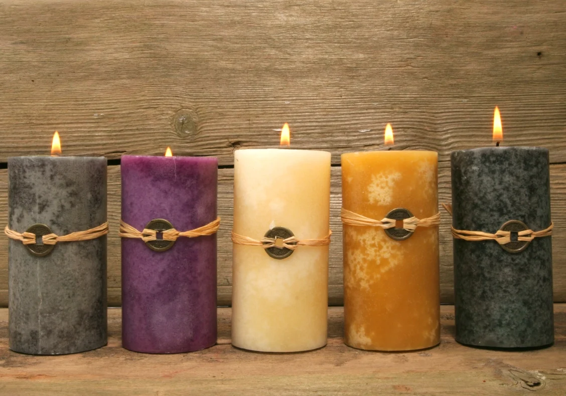 Five lit candles in gray, purple, cream, yellow, and dark gray stand in a row on a wooden surface. Each has a rustic twine and metal adornment. Background is a wooden panel.