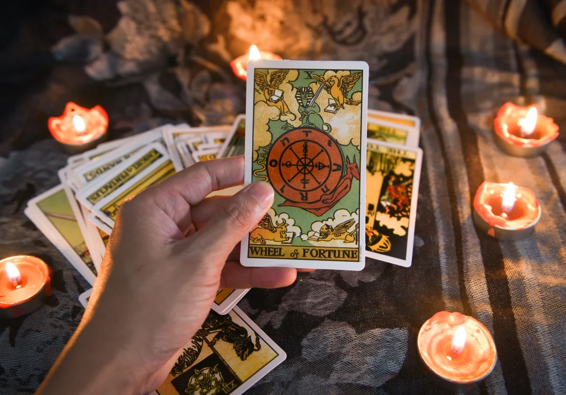 Hand holding a "Wheel of Fortune" tarot card surrounded by lit candles and other tarot cards on a dark surface.